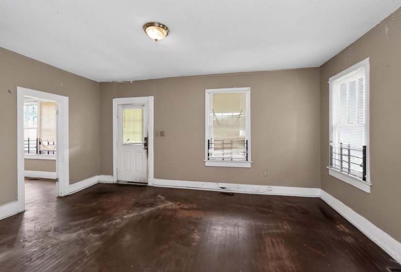 Spacious room featuring wood-finish flooring, neutral-toned walls, multiple windows with security bars, a white paneled door, and a flush mount ceiling light fixture