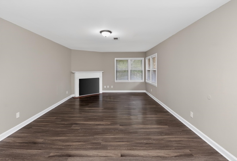Living area featuring dark wood-finish flooring, light neutral wall paint, white trim, a corner fireplace with a white mantel, and multiple windows for natural light