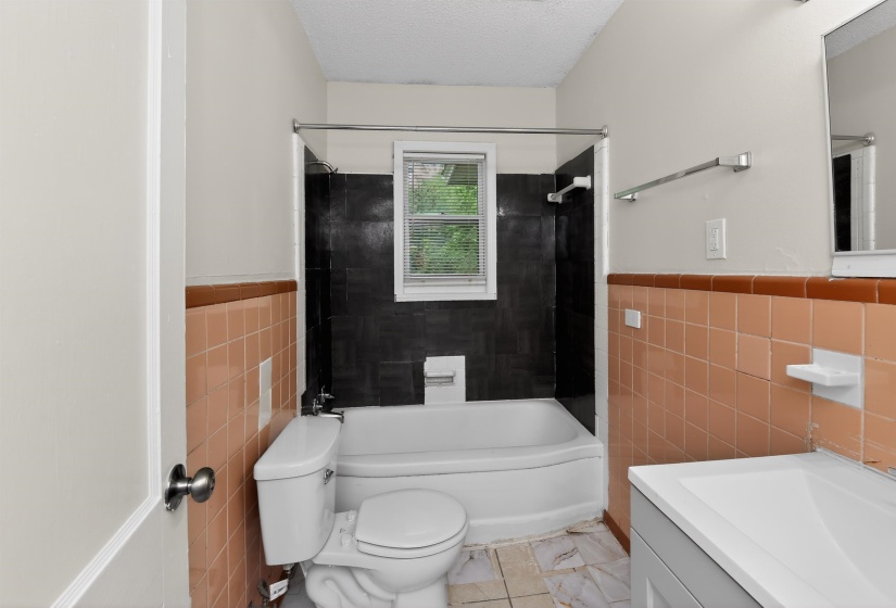 Bathroom featuring a white bathtub with a dark tiled surround, a window with blinds, and a toilet