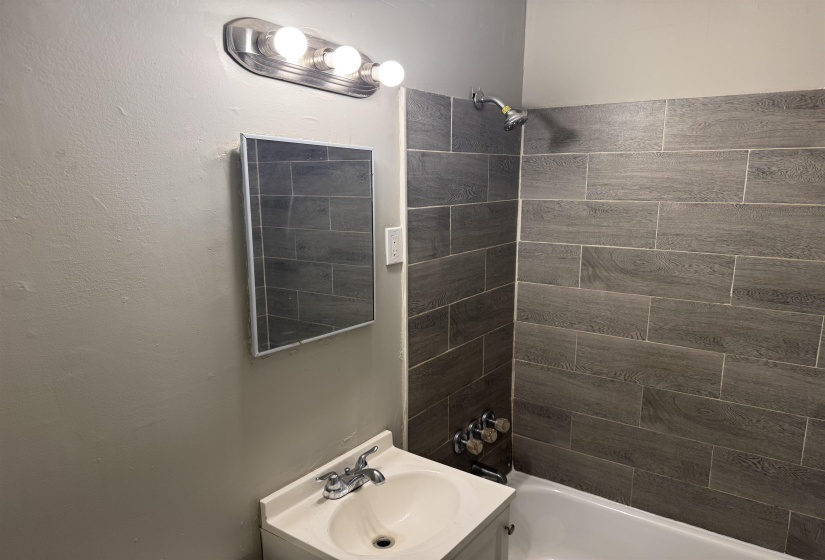 Bathroom featuring a built-in bathtub with dark gray horizontal tile surround, a wall-mounted shower head, and a vanity with an integrated sink and chrome faucet