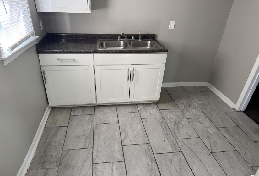 Kitchenette featuring white cabinetry with brushed nickel hardware, a stainless steel double basin sink, dark speckled countertop, and grey rectangular tile flooring