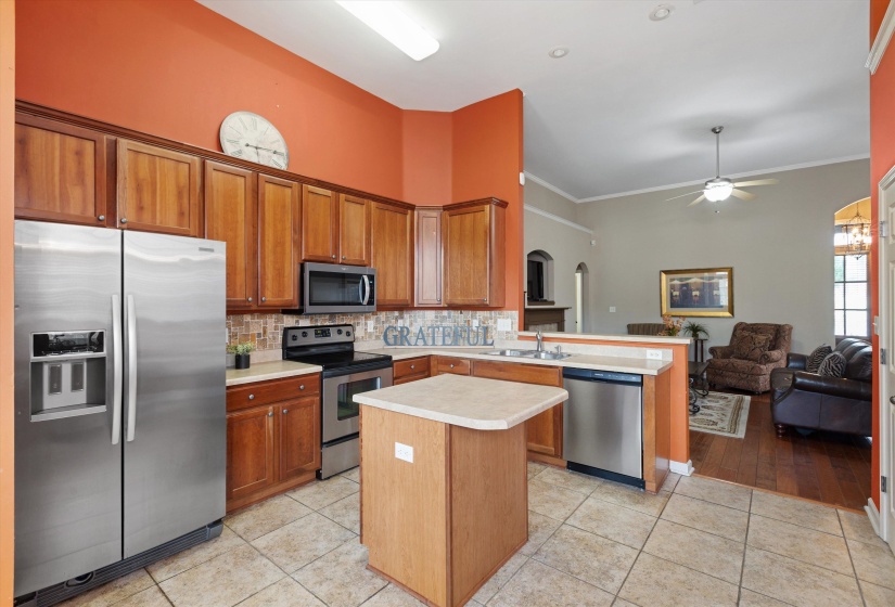 Open-concept kitchen featuring wood cabinetry, stainless steel appliances, a tiled backsplash, a central island, and tile flooring