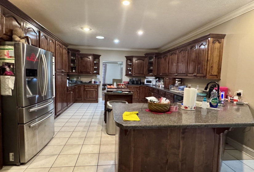 Kitchen featuring extensive dark wood cabinetry with crown molding, a stainless steel refrigerator, recessed lighting, tile flooring, and a central island with a stone-finish countertop
