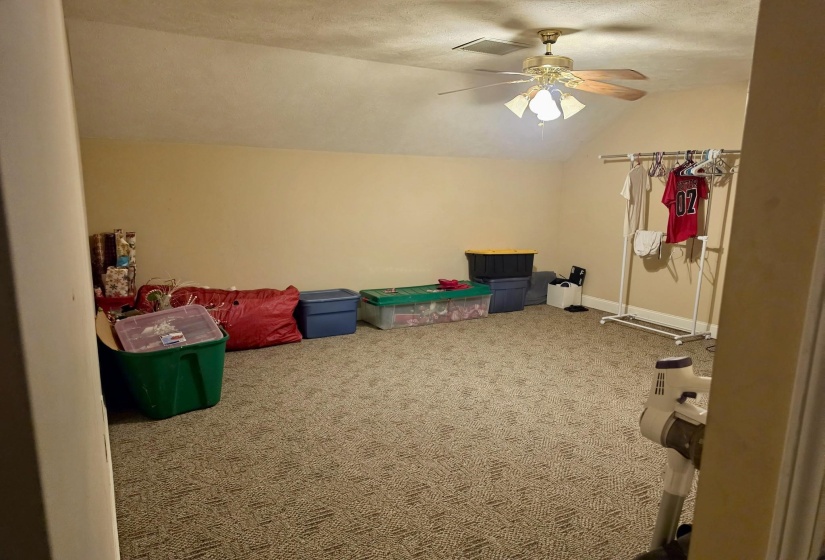 Spacious room featuring sloped ceilings, textured carpeting, a ceiling fan with integrated lighting, and neutral wall tones