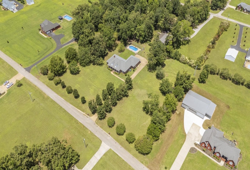 Sprawling property featuring a primary residence with a dark gray roof, a separate large outbuilding with a light gray roof, extensive green acreage, mature trees, and paved driveways