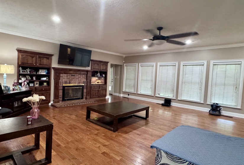 Spacious living area featuring hardwood flooring throughout, a brick fireplace with built-in wood cabinetry, a ceiling fan with integrated lighting, recessed lighting, and multiple windows with blinds