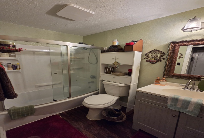 Bathroom featuring a single vanity with an integrated sink, a framed mirror, a toilet, a shower-tub combination with sliding glass doors, and wood-finish flooring