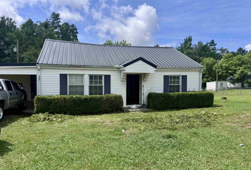 White siding exterior with a metal roof, featuring a covered carport, dark blue window shutters, and established front landscaping