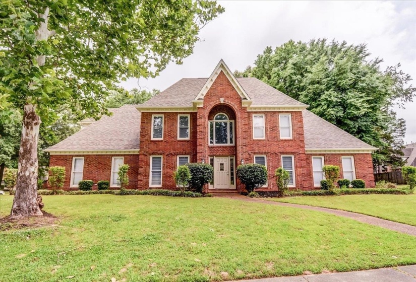 Traditional brick exterior featuring a prominent gabled entry with an arched window, white trim detailing, a concrete walkway, and a manicured lawn with mature trees