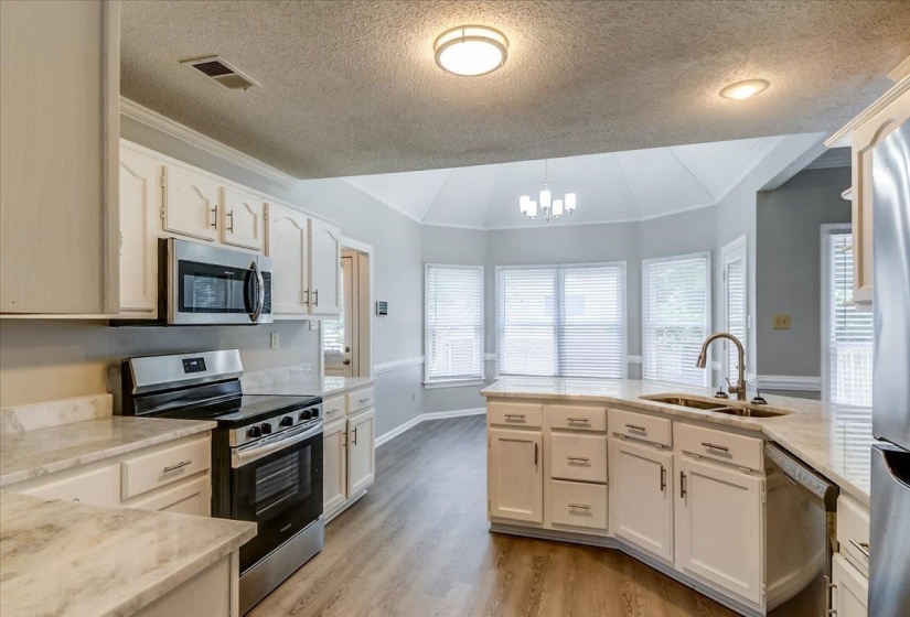 Kitchen featuring a central island with an integrated sink, stainless steel appliances, abundant white cabinetry, light-toned countertops, and wood-finish flooring