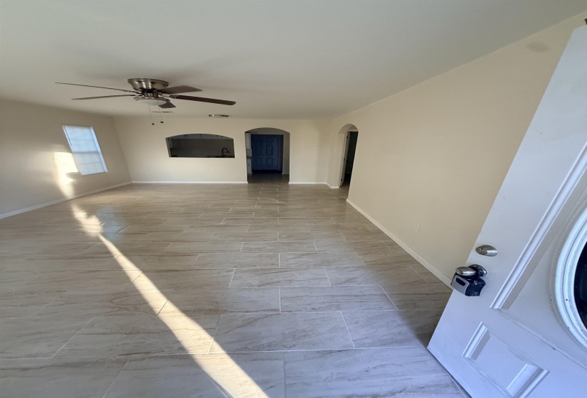 Spacious living area featuring neutral-toned walls, tile flooring, a ceiling fan with integrated lighting, a pass-through window with an arched top, and an arched doorway