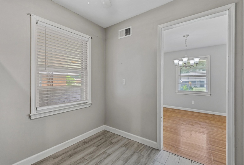 Interior space featuring light gray walls, wood-finish flooring, a window with blinds, and an open doorway revealing a second room with a chandelier and hardwood flooring