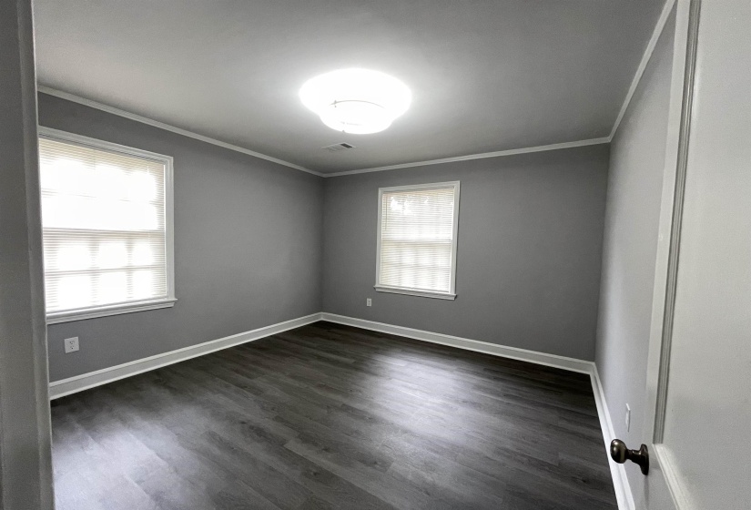Room featuring wood-finish flooring, two windows with blinds, a ceiling light fixture, crown molding, and gray wall paint