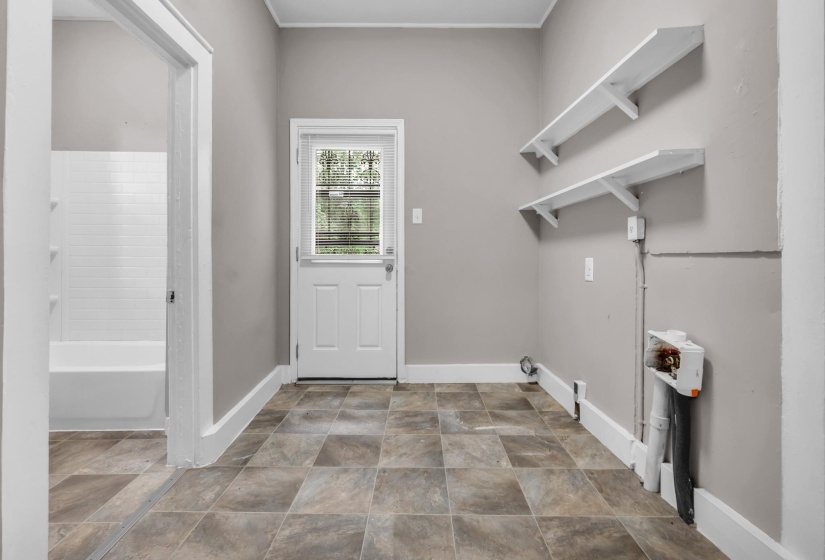 Utility area featuring square tile flooring, a white paneled door with glass insert, two wall-mounted shelves, and an opening to an adjacent room with a white bathtub