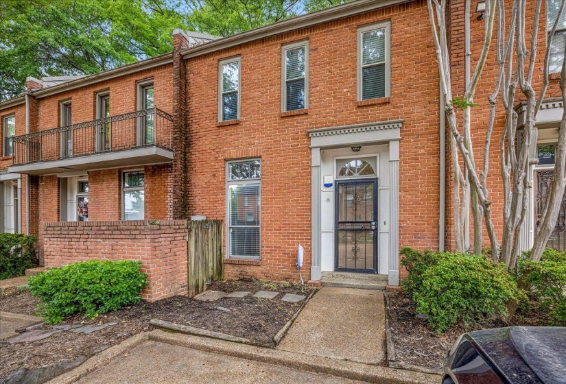 Brick facade townhome featuring a concrete walkway, mature shrubbery, a wrought iron security door, and a decorative balcony