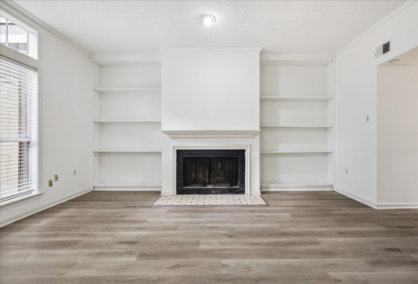 Living area featuring a fireplace with a white mantel, built-in shelving, wood-finish flooring, and a large window