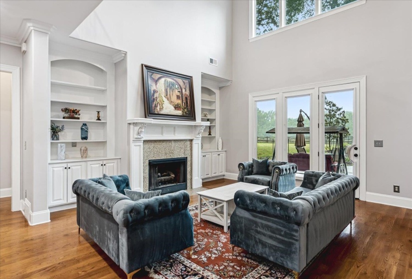 Living area featuring a soaring ceiling, wood-finish flooring, a fireplace with a decorative mantel and stone surround, built-in shelving with lower cabinetry, and expansive glass doors opening to the exterior