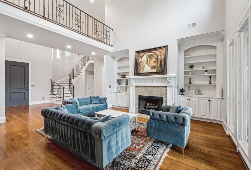 Spacious living area featuring high ceilings, hardwood flooring, an ornate fireplace with built-in cabinetry, large windows, and a prominent staircase with wrought iron balusters