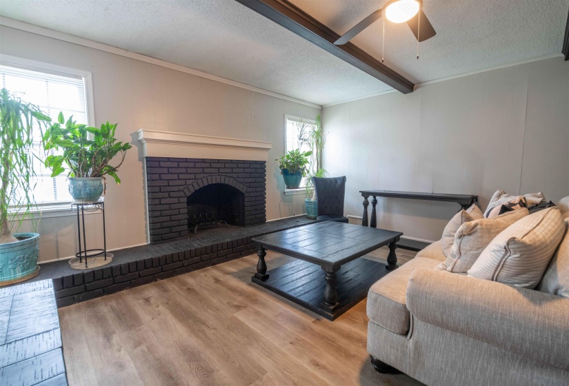 Living area featuring a painted brick fireplace with a white mantel, light wood-finish flooring, and a ceiling fan with dark wood-look beams
