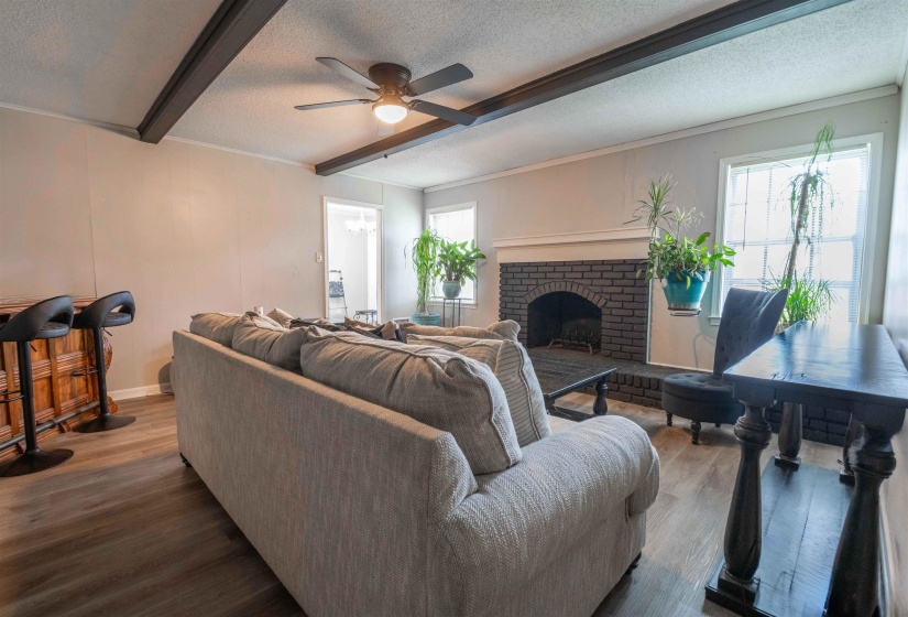 Living area featuring wood-finish flooring, exposed ceiling beams, a brick fireplace with a white mantel, and multiple windows