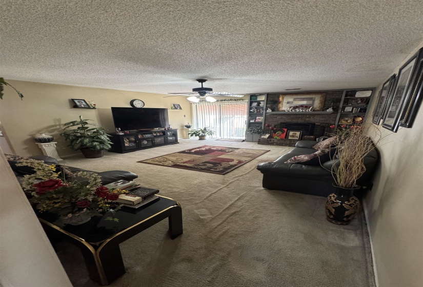 Living area featuring neutral-toned carpeting, a ceiling fan with integrated lighting, and a brick fireplace with built-in shelving