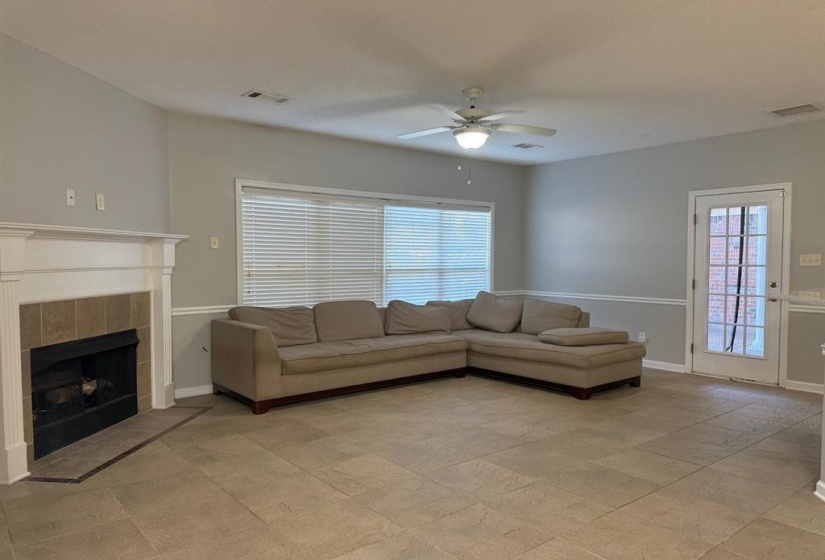 Spacious living area featuring neutral-toned tile flooring, a white mantel fireplace with tile surround, a ceiling fan with integrated lighting, and multiple windows with blinds