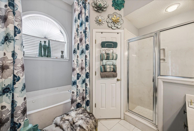 Spacious bathroom featuring a jetted tub with an arched window, a separate glass-enclosed shower, tile flooring, and overhead recessed lighting