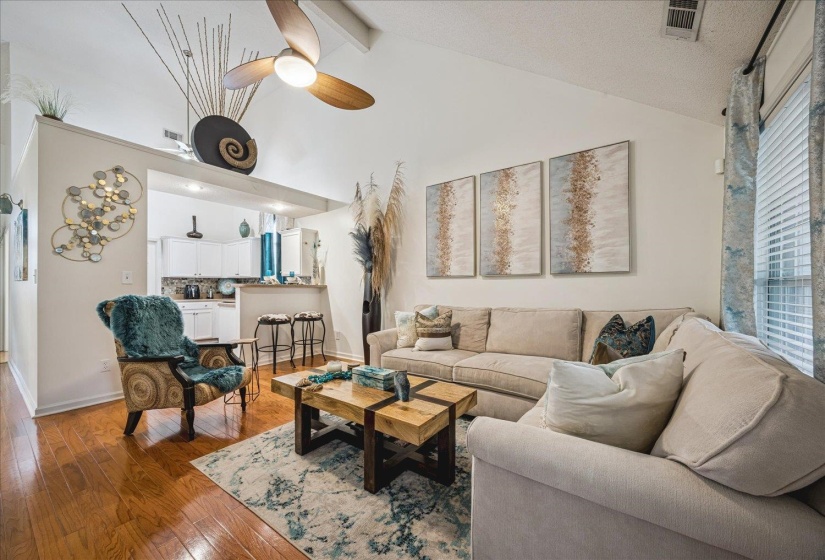 Vaulted ceiling with a ceiling fan, hardwood flooring, and an open concept layout to the kitchen featuring a breakfast bar, white cabinetry, and a tile backsplash
