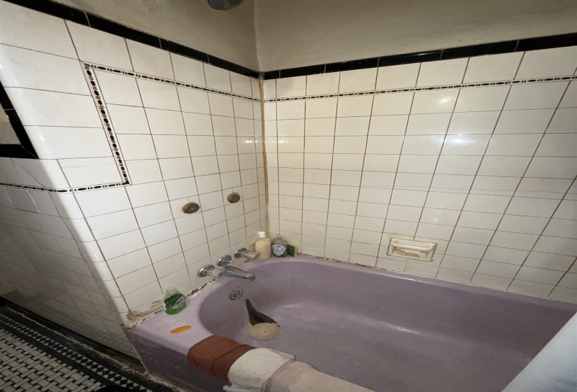 Enclosed bathtub featuring an integrated shower head and wall-mounted faucet, surrounded by white square tile walls with black trim accents