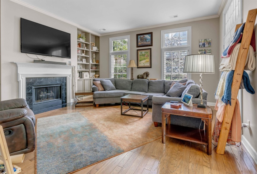 Living space featuring wood-finish flooring, a fireplace with a dark stone surround and white mantel, built-in shelving, and multiple windows