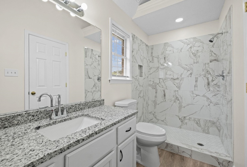 Bathroom featuring a walk-in shower with marble-look tile surround and built-in niche, white vanity with granite countertop and undermount sink, and wood-finish flooring