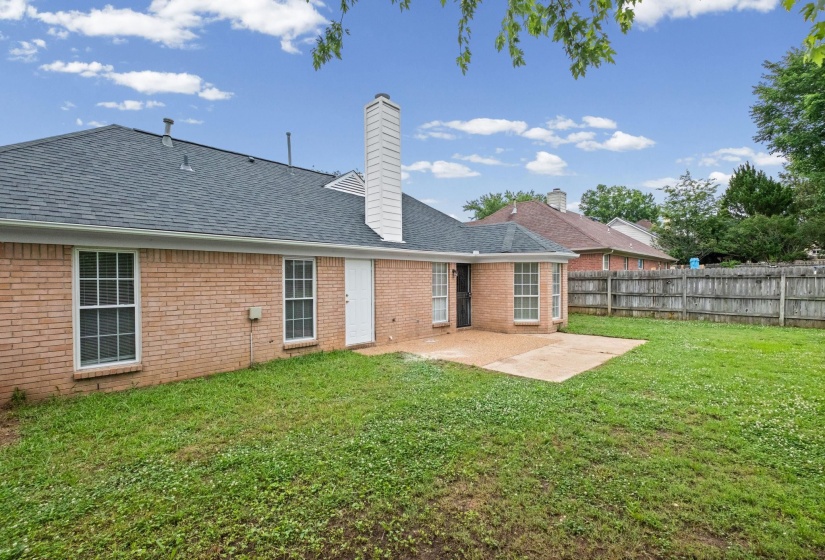 Expansive backyard featuring a concrete patio, brick exterior, dark shingle roof, white trim windows, and a wood privacy fence