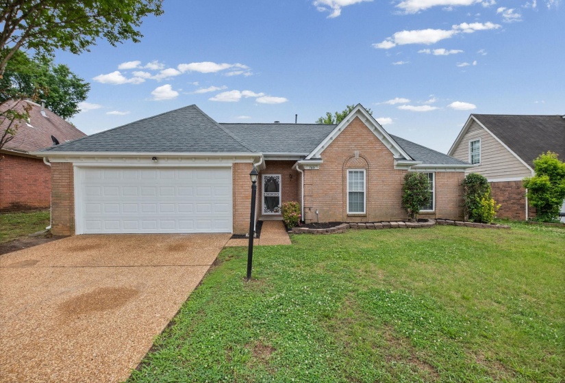 Brick exterior featuring a two-car garage with a white panel door, an exposed aggregate driveway, and a front yard with established landscaping
