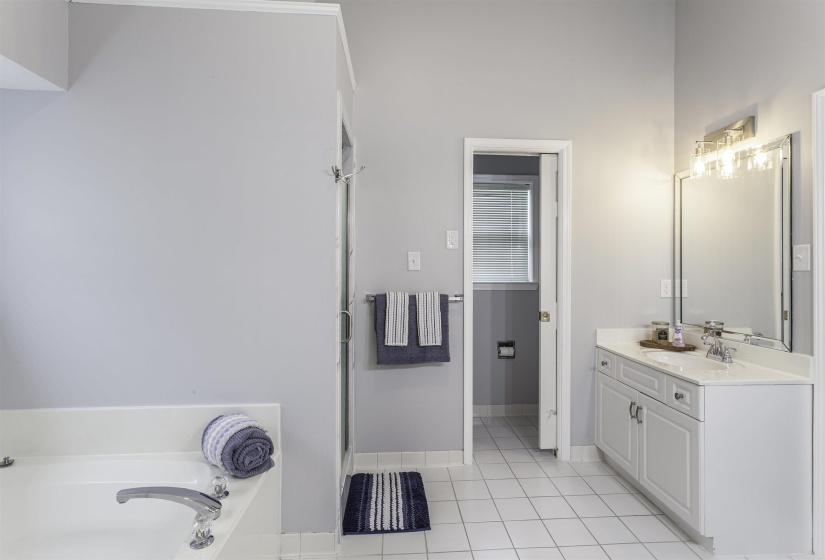 Bathroom featuring a built-in tub, separate shower with glass enclosure, white tile flooring, double vanities with integrated sink and chrome faucet, and a wall-mounted rectangular mirror with a four-light fixture