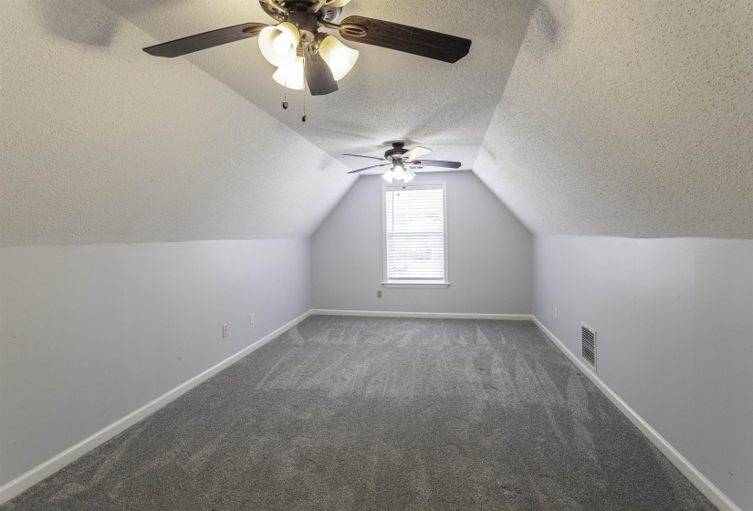 Spacious room featuring vaulted ceilings, two ceiling fans with integrated lighting, window with blinds, and neutral tone carpeting