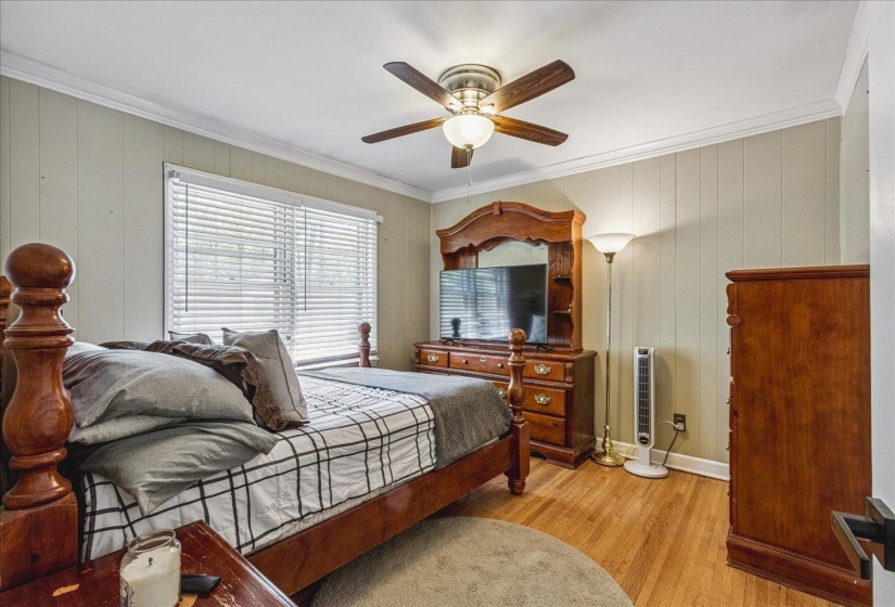 Bedroom featuring horizontal paneling walls, crown molding, a ceiling fan with light fixture, wood-finish flooring, and a window with blinds