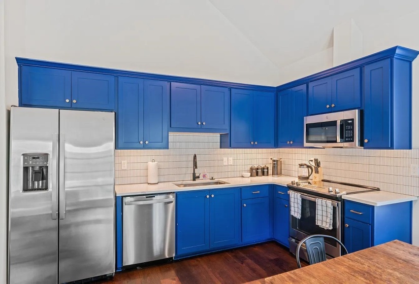 Kitchen featuring stainless steel appliances, blue cabinetry, dark wood finished floors, light stone counters, and vaulted ceiling