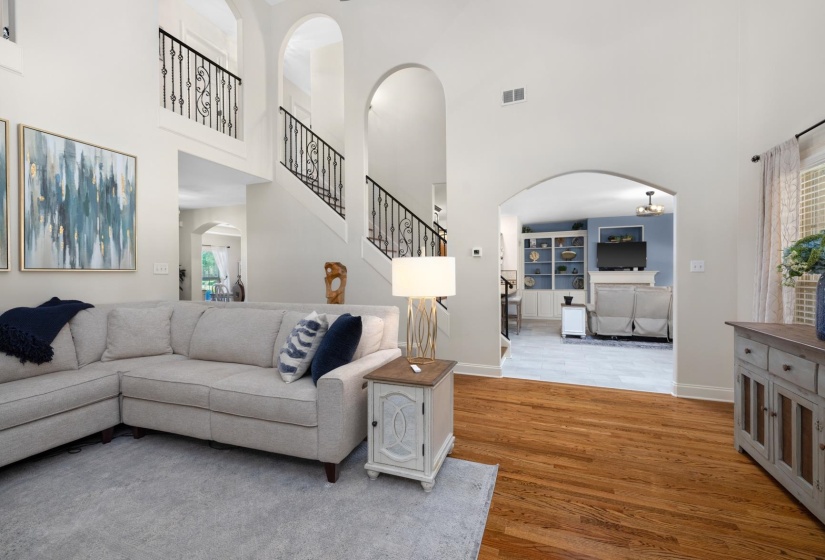 Expansive living area featuring hardwood flooring, arched doorways, a dual-story ceiling, a grand staircase with wrought iron balusters, and a recessed room with built-in shelving