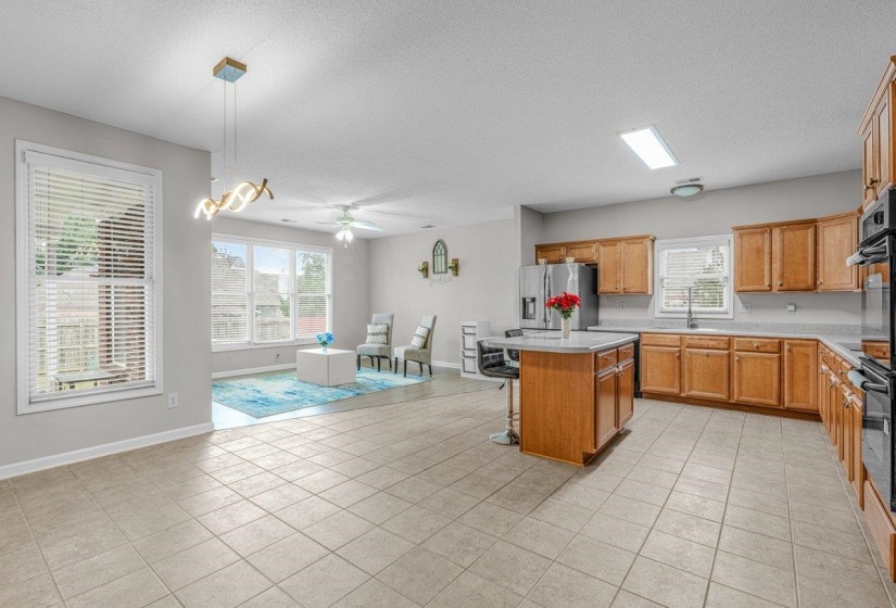 Spacious open-concept interior featuring light tile flooring, an expansive kitchen with wood cabinetry and a central island, stainless steel appliances, and a bright living area with multiple windows