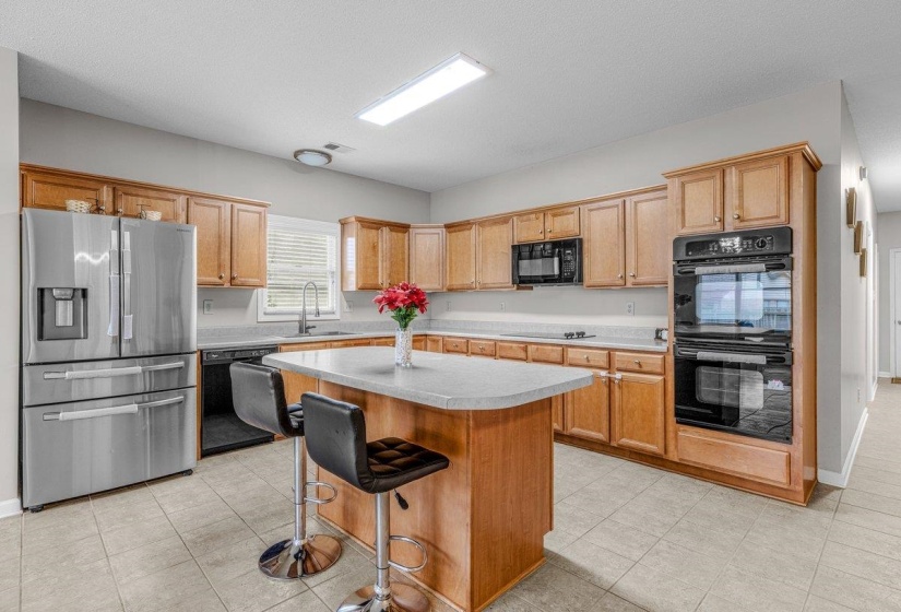 Kitchen featuring a central island with a light-toned countertop, stainless steel refrigerator, built-in double ovens, wood-finish cabinetry, and tiled flooring