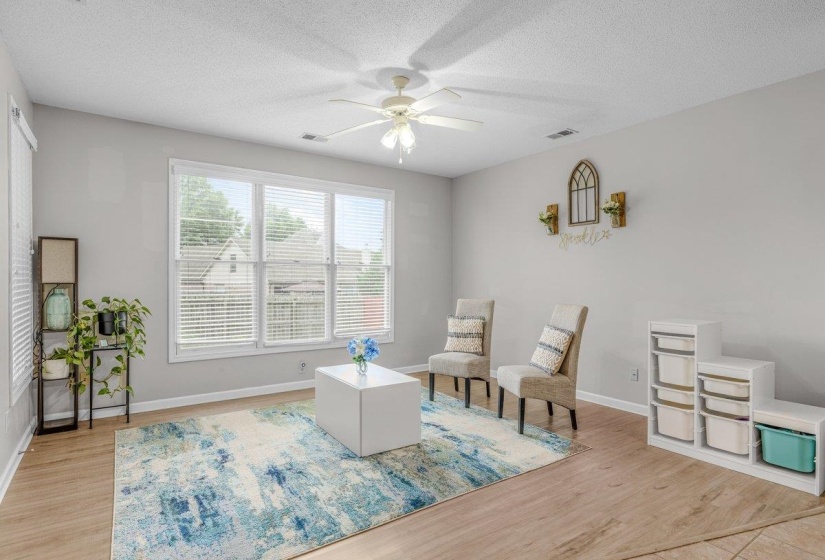 Spacious living area featuring light wood-finish flooring, a ceiling fan with integrated lighting, and multiple windows with blinds