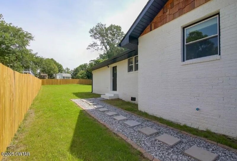 Expansive fenced backyard featuring a manicured lawn, a gravel and paver pathway, and a white brick exterior