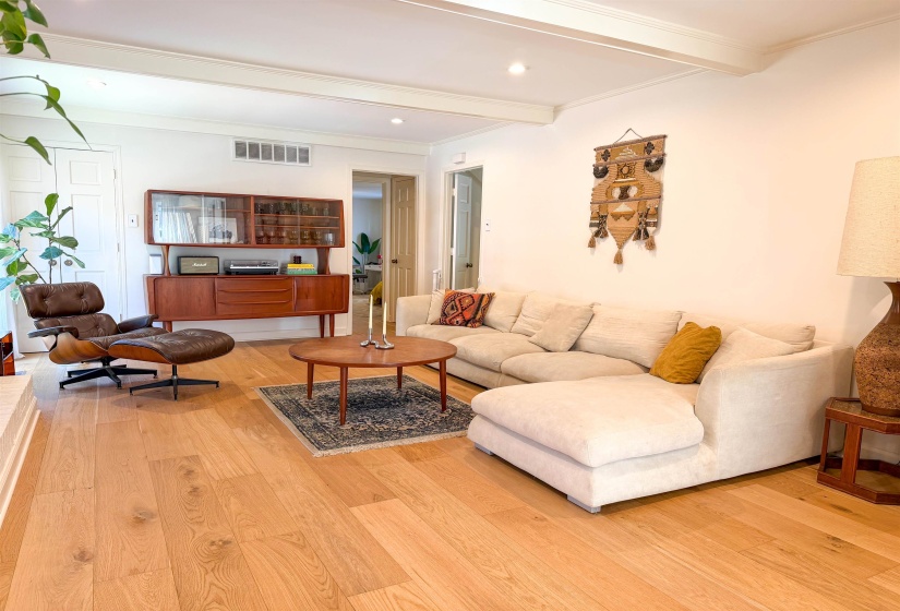 Spacious living area featuring light wood-finish flooring, white walls, and exposed ceiling beams