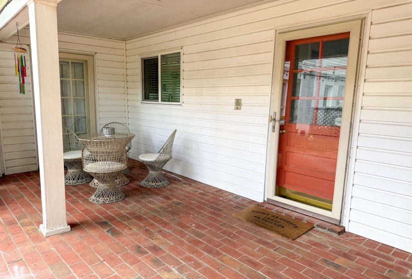 Covered porch featuring brick flooring, white horizontal siding, a red entry door with glass panels, a second entry door, and a window with blinds