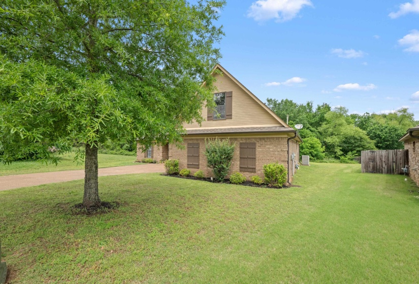Brick and siding exterior featuring a prominent tree, expansive green lawn, and a partial wooden fence