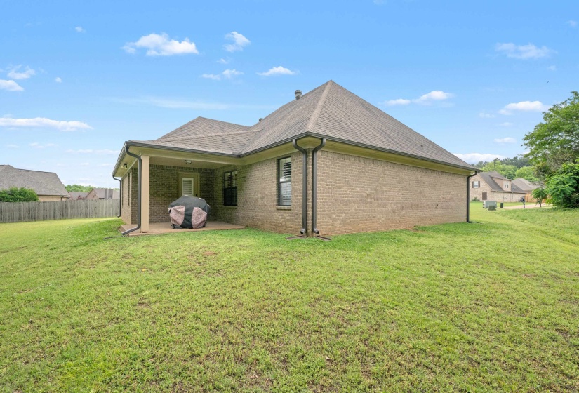 Brick exterior with a covered patio and concrete slab, featuring a shingle roof, dark gutters, and a grassy lawn