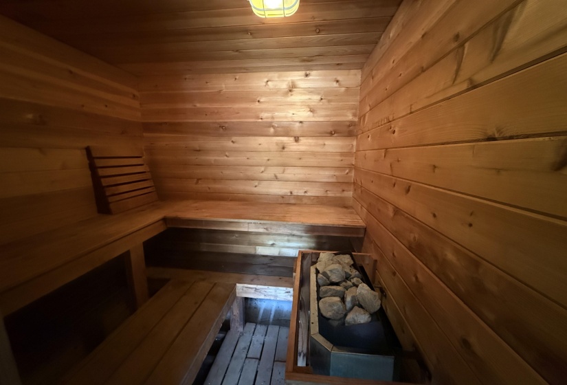 Wood-paneled sauna featuring a built-in heater with rocks, slatted wood benches, and a wall-mounted light fixture