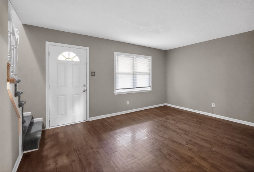Spacious living area featuring wood-finish flooring, neutral wall tones, and a bright window with blinds