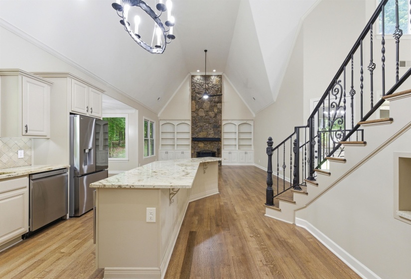 Vaulted ceilings highlight this open-concept interior featuring wood-finish flooring, a kitchen with light-toned cabinetry and stainless steel appliances, a stone-front fireplace with built-in shelving, and a staircase with ornate wrought iron balusters