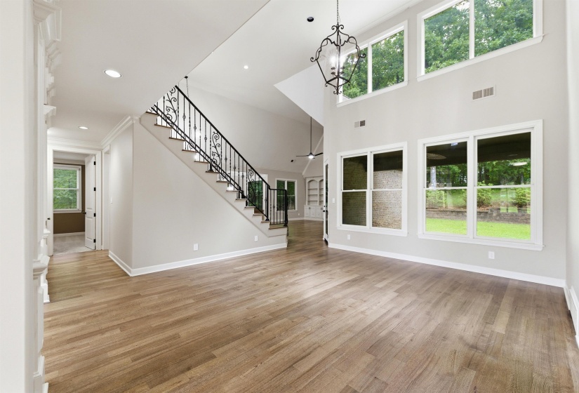 Open-concept living area with soaring ceilings, wood-finish flooring, a grand staircase with wrought iron balusters, a decorative chandelier, and extensive windows providing abundant natural light